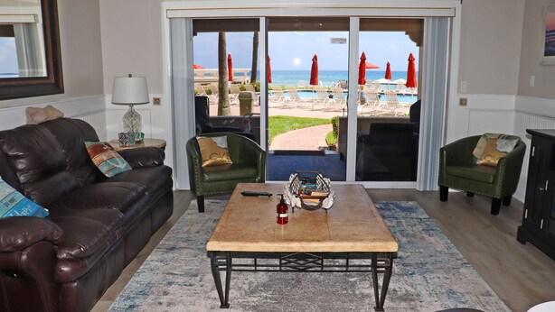 A living room with a leather sofa, two armchairs, a coffee table, and a view of a pool and beach through glass doors. - Steps to south pool deck, pool and ocean from this ground floor, three bedroom, three bath sleeps 8. (New Smyrna Beach)