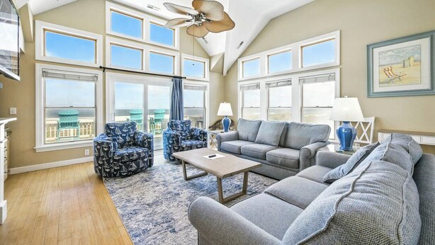 A spacious living room with a high ceiling, large windows, and a ceiling fan. There are two sofas, a coffee table, and patterned chairs. - Tipsy Mermaid | Oceanfront | Private Pool, Hot Tub | Nags Head (Nags Head)