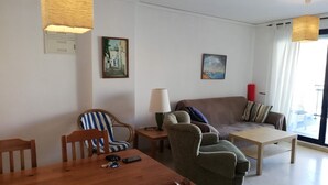 A living room with a wooden dining table, two armchairs, a sofa, and a lamp. There are paintings on the walls and a window with curtains. - Riumar BL. b 1º SOLO FAMILIAS (Xeraco Playa)