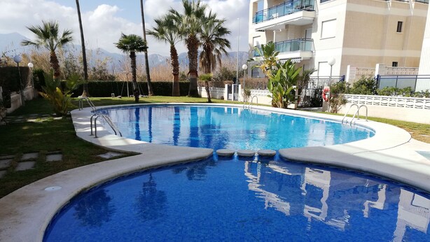 A swimming pool with a curved edge, surrounded by palm trees and a building with balconies. - Riumar BL. b 1º SOLO FAMILIAS (Xeraco Playa)