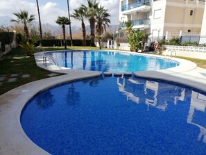 A swimming pool with a curved edge, surrounded by palm trees and a building with balconies.