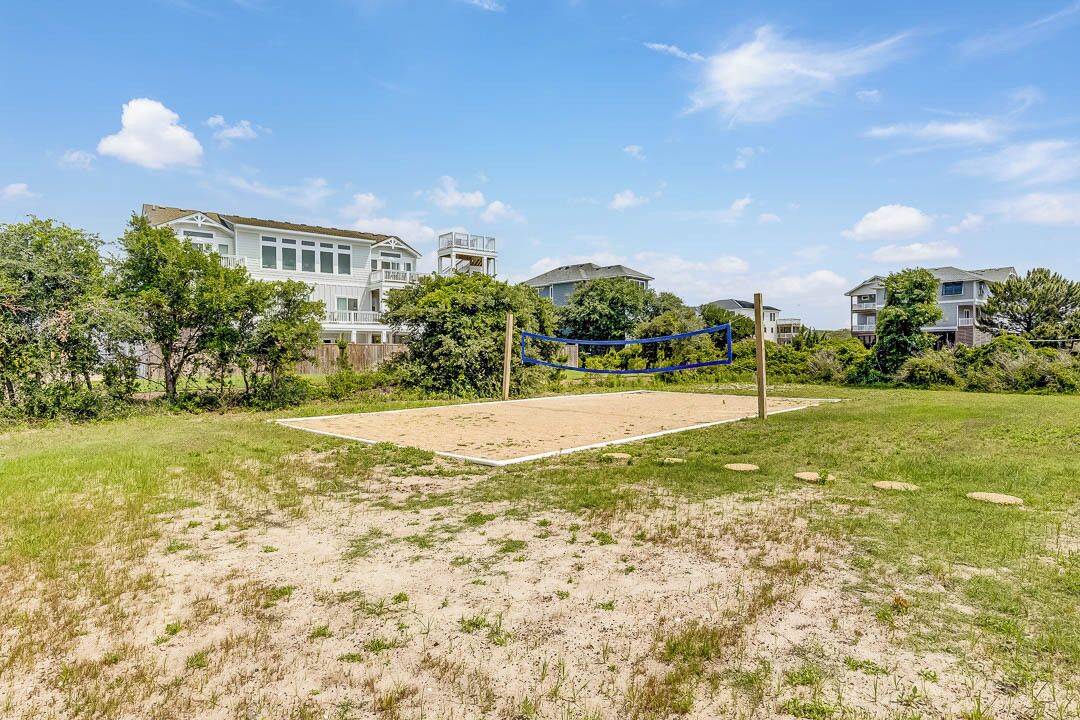 Una cancha de voleibol con red, rodeada de césped y árboles, con casas al fondo.