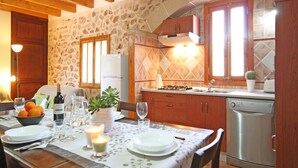 A rustic kitchen with stone walls, wooden beams, and a tiled backsplash. There is a dining table set with plates, glasses, and a candle. A refrigerator, stove, and dishwasher are visible. - Historic finca with modern comfort and panoramic views, pool, calm nature, WiFi (Son Serra de Marina)