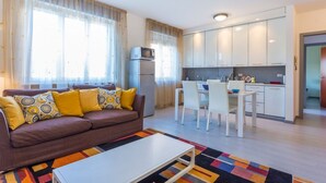 Un salon comprenant un canapé, une table basse et un coin repas avec des chaises et une table. On aperçoit une cuisine avec des placards et des appareils électroménagers à l’arrière-plan.
