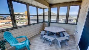 A covered balcony with a wooden table and chairs, a turquoise chair, and large windows offering a view of buildings and a parking lot.