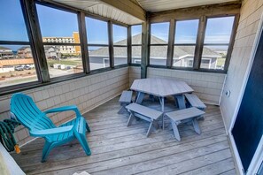 Un balcón techado con una mesa y sillas de madera, una silla turquesa y amplias ventanas que dan a vistas de edificios y un estacionamiento.