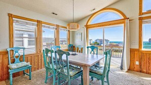 A dining area with a wooden table and six chairs, large windows with wooden frames, and a view of the ocean.