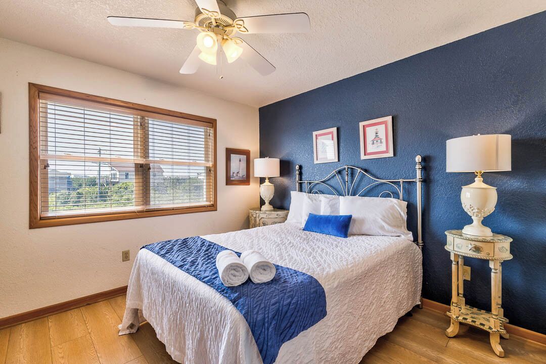 Une chambre à coucher comprenant un lit, un ventilateur de plafond, une fenêtre avec des stores, deux tables de chevet avec des lampes et deux tableaux encadrés accrochés au mur.