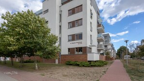Un edificio moderno de varios pisos con base de ladrillo y pisos superiores blancos, que cuenta con balcones y un carril bici en la parte delantera.
