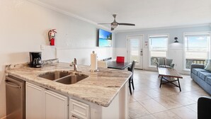 A kitchen with a marble countertop, a coffee maker, and a sink. There is a ceiling fan, a fire extinguisher, and a window with a view of the beach. - Beachfront Escape | Ocean Views, Heated Pool & Hot Tub (Corpus Christi)