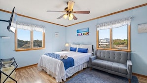 A bedroom with a bed, a grey sofa, a ceiling fan, and large windows with a view of the landscape. - Even Steven | 570 ft from the beach | Private Pool | Duck (Duck)