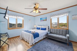 Une chambre à coucher avec un lit, un canapé gris, un ventilateur de plafond et de grandes fenêtres donnant sur le paysage.