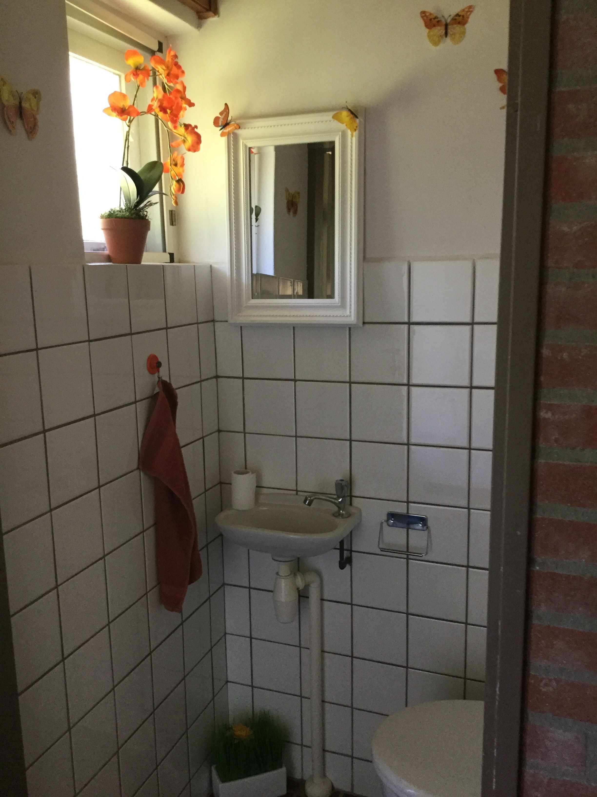 Ett badrum med vita kakelbeklädda väggar, ett handfat, en spegel och ett fönster med en krukväxt.