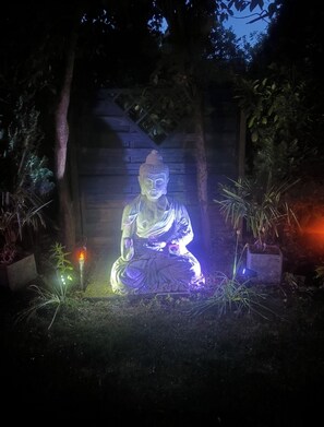 A statue of a seated figure in a garden setting with surrounding vegetation and illuminated by artificial lighting.