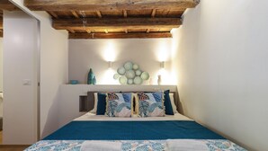 Um quarto com teto de madeira, cama com colcha azul e branca estampada, duas almofadas com desenho de folha e duas luminárias fixadas na parede.