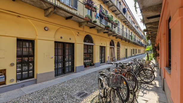 Ruas de pedra com edifícios amarelos, bicicletas estacionadas ao longo da calçada e varandas com plantas pendentes.
