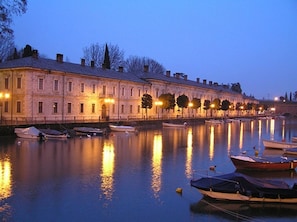 A waterfront hotel with a row of rooms along the river, boats docked in the water, and street lamps along the walkway.