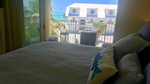 A hotel room with a bed, a view of a building with blue and white architecture, and a balcony with a railing. - Key Colony Beach 3/3 Townhouse - Ocean View (Key Colony Beach)