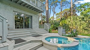 Un área junto a la piscina con un jacuzzi, escalones que conducen a una terraza cubierta y exuberante vegetación tropical.