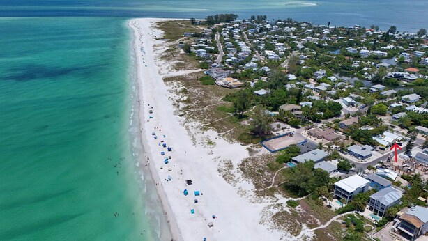 Vista aérea de un pueblo costero con una larga playa de arena, aguas turquesas cristalinas y un grupo denso de casas y edificios.