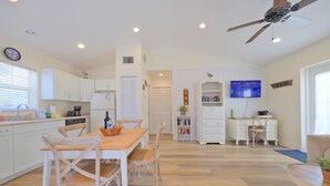 Una cocina amplia con armarios blancos, una mesa de comedor con sillas, un ventilador de techo y un televisor montado en la pared.