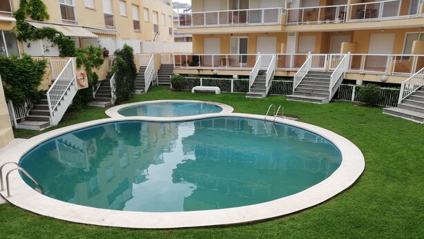 A swimming pool with a surrounding grassy area and a multi-story building in the background. - PARQUEMAR VIII BL. II 1º SOLO FAMILIAS! (Xeraco Playa)