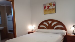 A neatly made bed with white linens and a folded towel, a wooden headboard, bedside lamps, and a wall-mounted painting of flowers. - PARQUEMAR VIII BL. II 1º SOLO FAMILIAS! (Xeraco Playa)