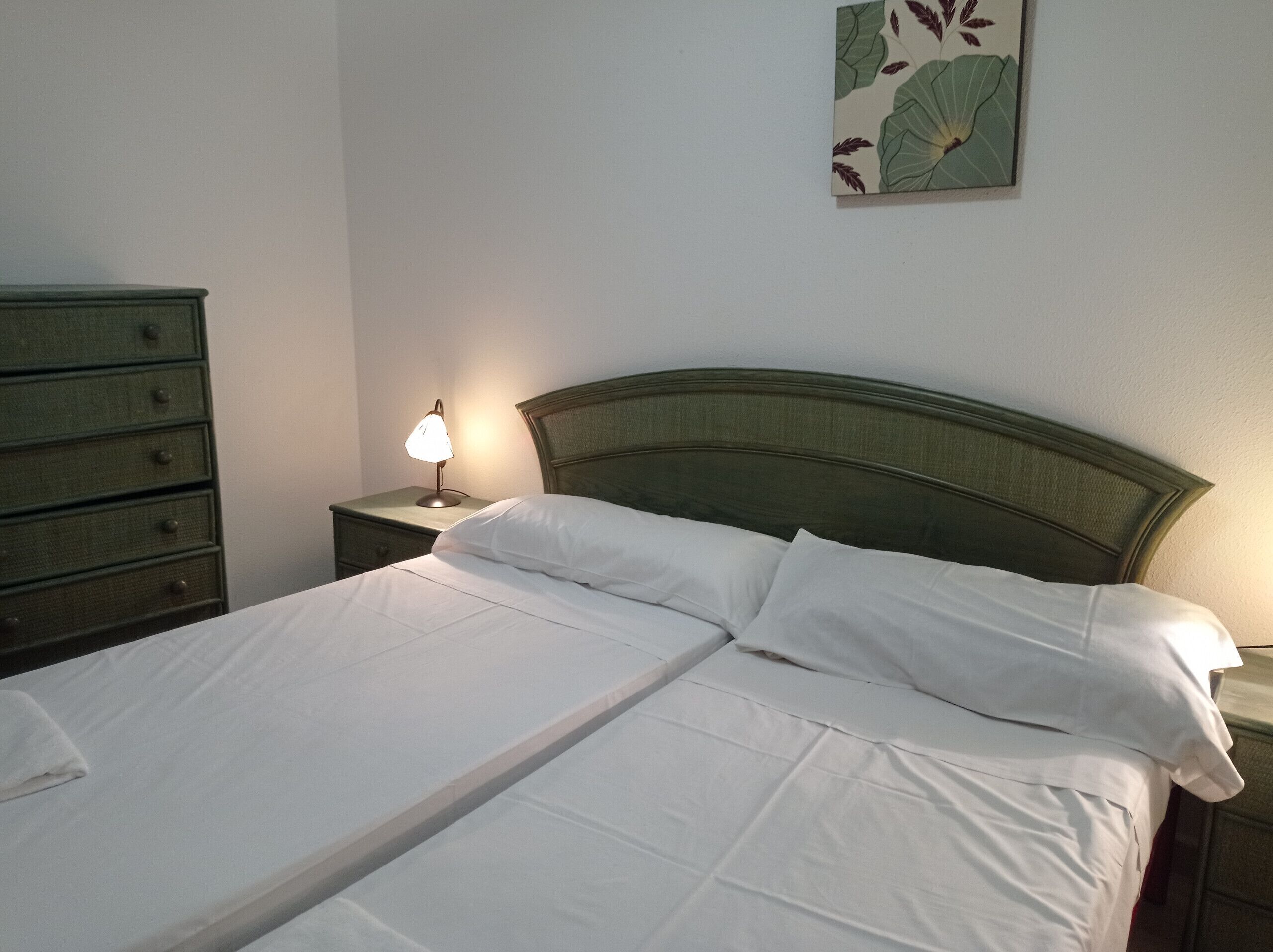 A double bed with white linens, a nightstand with a lamp, and a dresser with multiple drawers.