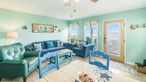 A living room with teal walls, blue furniture, a ceiling fan, and a view through the door. - Beachfront Escape | Pool, Hot Tub & Ocean Views (Corpus Christi)