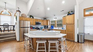Uma cozinha moderna com armários de madeira, uma geladeira de aço inoxidável, uma ilha embutida com banquetas e uma área de jantar com mesa e cadeiras.