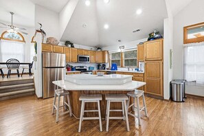A modern kitchen with wooden cabinets, a stainless steel refrigerator, a built-in island with stools, and a dining area with a table and chairs.