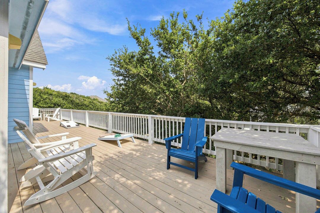 A wooden deck with outdoor furniture, including white Adirondack chairs and a blue chair, surrounded by a white fence and trees.