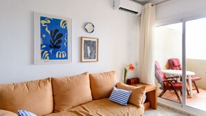 A modern living room with a beige sofa, a small table, and a view of a balcony with red chairs.