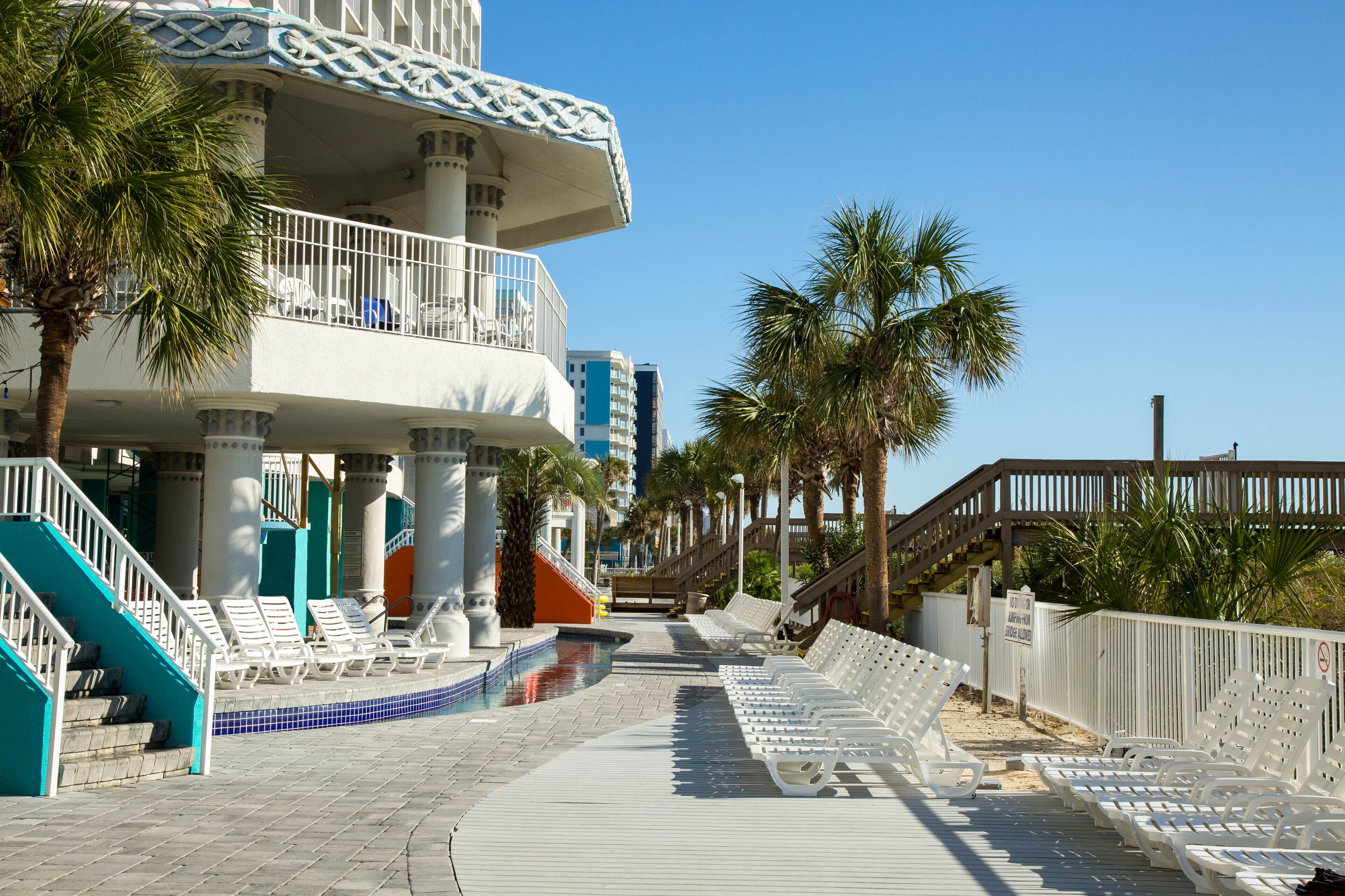 A resort with a pool, lounge chairs, palm trees, and a staircase leading to a balcony.