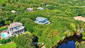 Une grande maison à plusieurs étages, entourée d’une végétation luxuriante et bordée d’un petit étang.