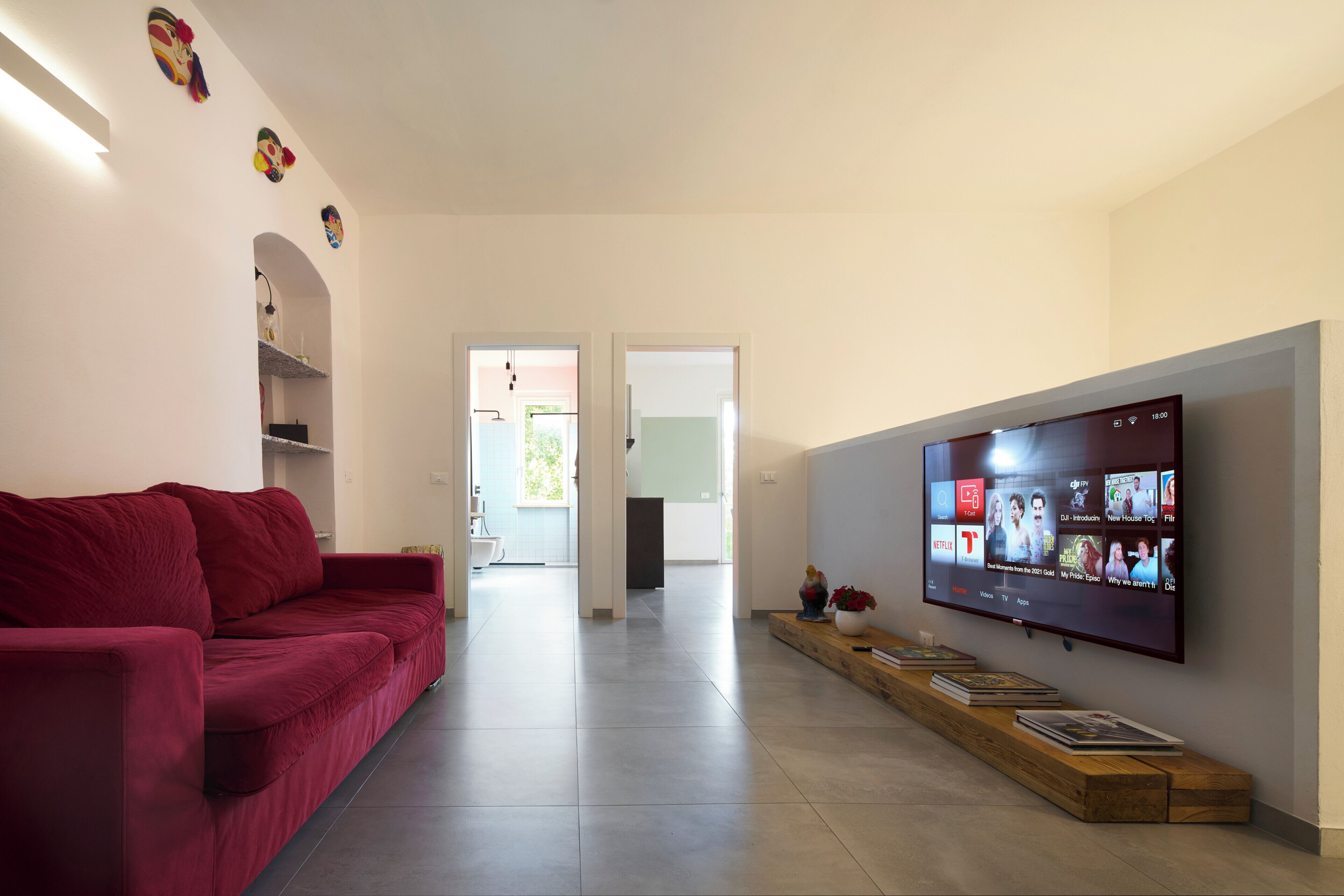 Un salon moderne avec un canapé rouge, un téléviseur à écran plat fixé au mur et une table d’appoint en bois sur laquelle sont posés des livres.