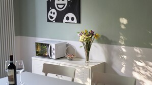 Un comedor con una mesa blanca, un microondas, un florero con flores y una botella de vino.