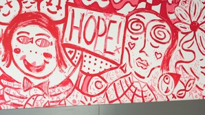 Una habitación con una cama, un bote de basura rojo y una pared con grafitis rojos.
