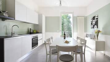 Una cucina moderna con mobili bianchi, un tavolo da pranzo con sedie, un bollitore, una bottiglia di vino e una finestra vista alberi.