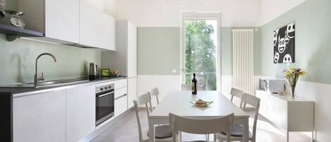 A modern kitchen with white cabinetry, a dining table with chairs, a kettle, a wine bottle, and a window with a view of trees.
