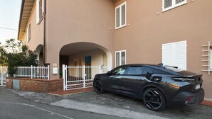 Un coche negro estacionado frente a un edificio color crema con contraventanas blancas y una valla blanca.