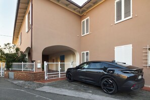 Un coche negro estacionado frente a un edificio color crema con contraventanas blancas y una valla blanca.