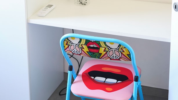 Un escritorio blanco con una lámpara, una silla de diseño colorido y una pared decorada con caritas sonrientes.