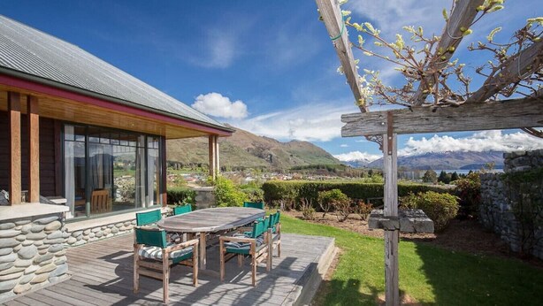 A wooden deck with a table and chairs, a stone wall, and a pergola with climbing plants. - Bullock Creek Chalet · Lake & mountain views (Wanaka)