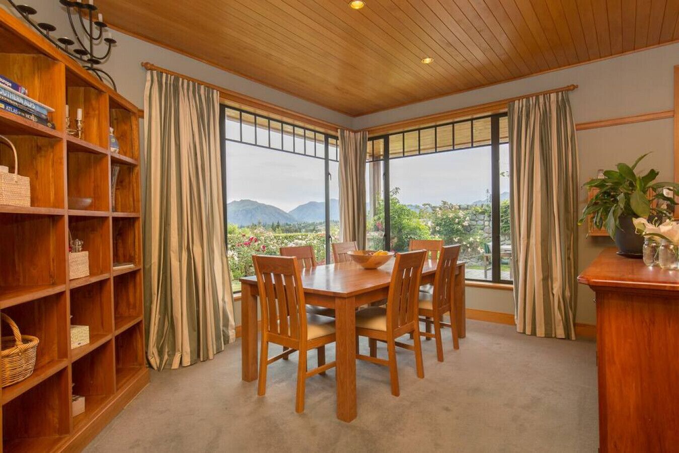 A dining area with a wooden table and chairs, large windows with curtains, and a bookshelf with various items.