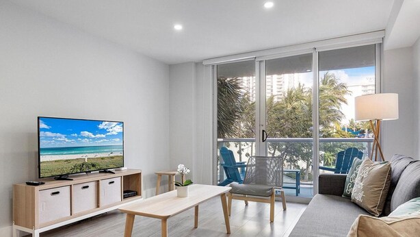 A modern living room with a flat-screen TV, a sofa, a dining table, and a balcony with a view of palm trees and buildings. - Family-Friends Getaway- Pool, Beach & Parking free (Sunny Isles Beach)
