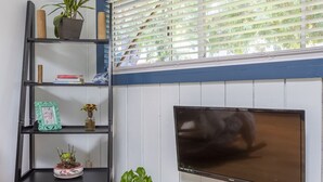 A room with a wooden floor, a black shelving unit with plants, a mounted TV, and a window with blinds. - OCEAN BEACH, #7 ZEN, RETRO COTTAGE STUDIO, ONLY 3 BLOCKS TO THE BEACH 🏖 (San Diego)