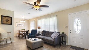 A living room with a ceiling fan, a dining table with chairs, a sofa, an ottoman, and a door with a stained glass window.