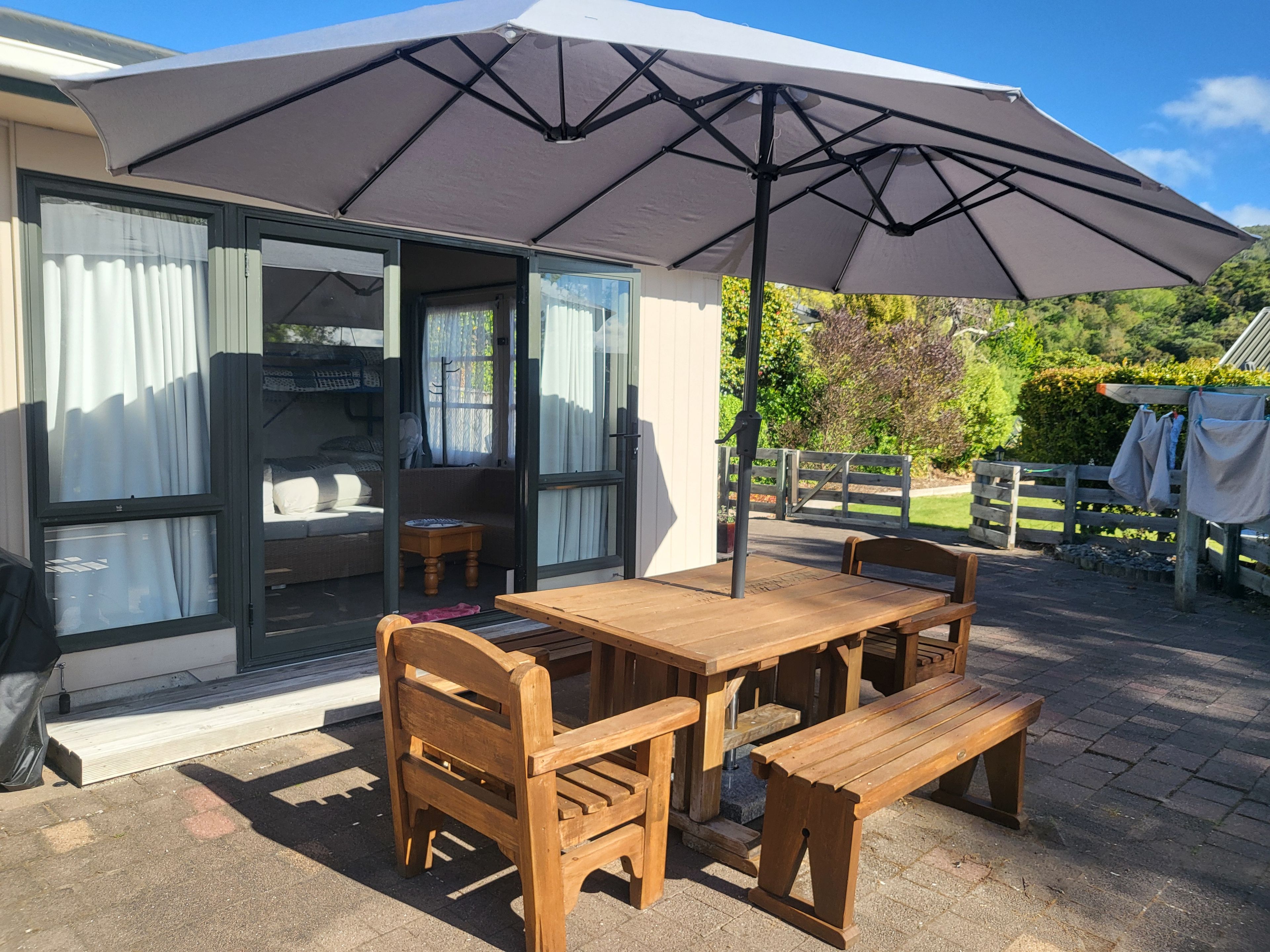 Une terrasse extérieure avec une table et des chaises en bois, un grand parasol et une porte-fenêtre coulissante donnant sur une pièce comprenant un canapé et une petite table.
