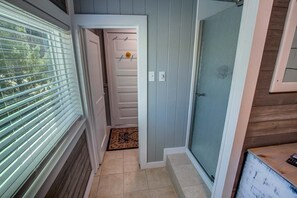 Un pasillo con una puerta que da a una habitación, una ventana con persianas y una alfombra pequeña en el suelo.